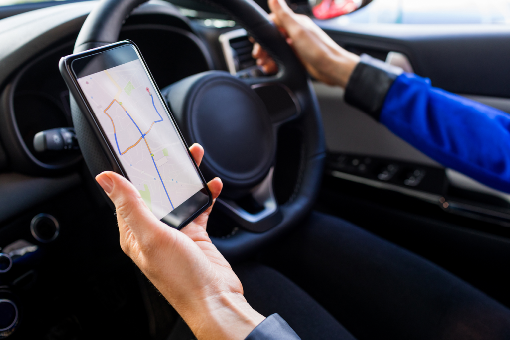 Driver holding a smartphone with a car tracker app showing live GPS route, dashboard and steering wheel visible in the background
