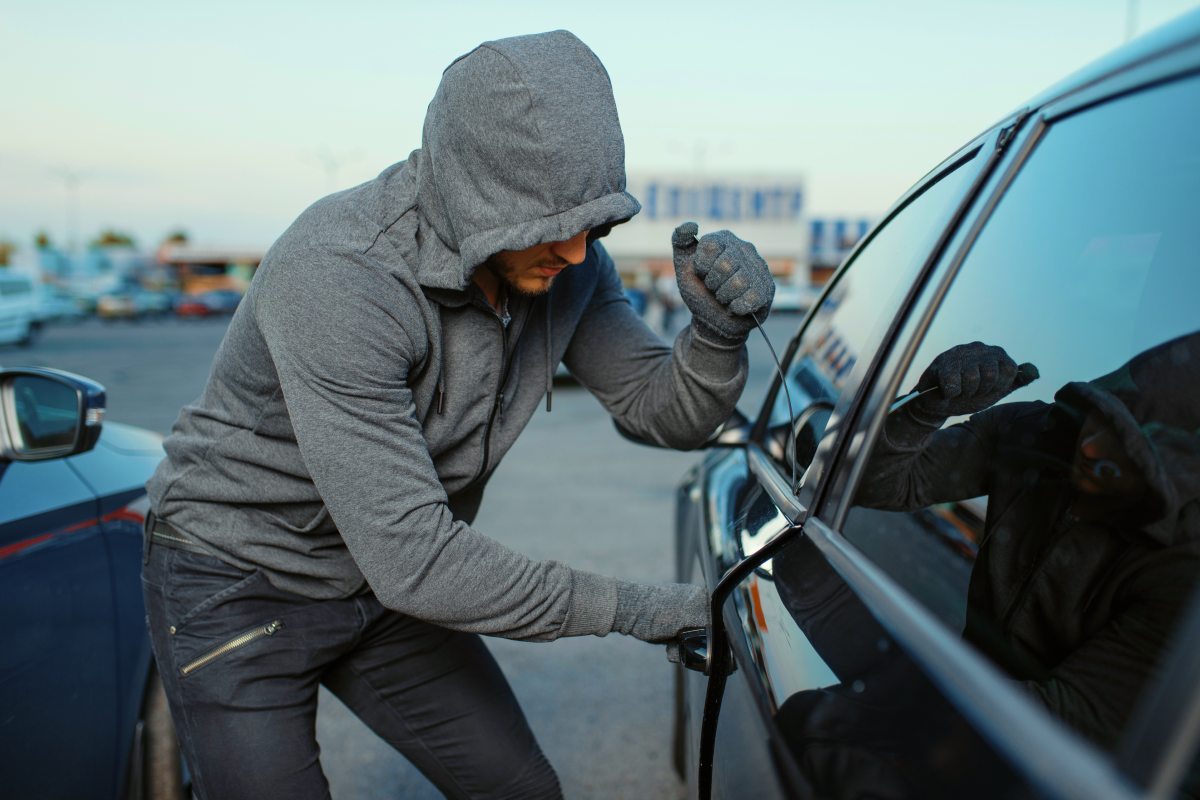 Hooded thief using a slim jim tool to break into a car door handle, illustrating the physical break-ins that keyless car theft prevention measures like CAN bus immobilisers don't need to worry about